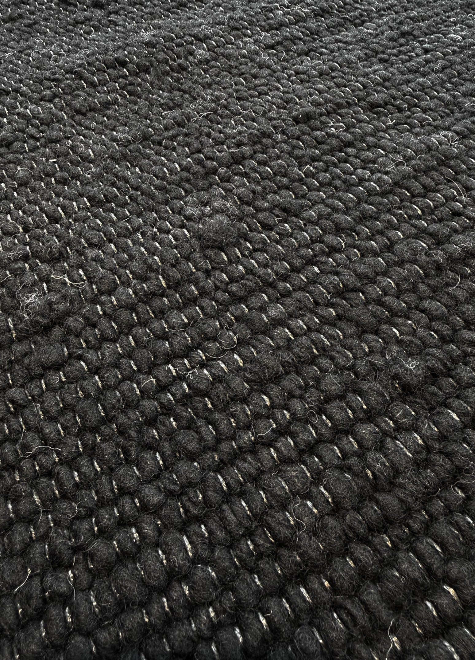 Natūralios vilnos kilimas su smulkiu bangelės raštu, kuris matosi iš arti. Kilimo spalva yra juoda ir tamsiai pilka.
