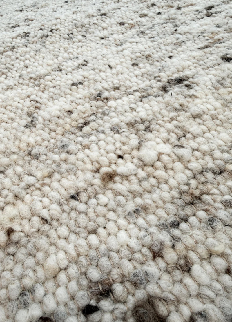 Mellow Sand kilimas iš arti, kur matoma jo tekstūra su smulkiais vilnos rutuliukais ir smėline, pilka, juoda spalva ir šviesiai rudais siūlais.